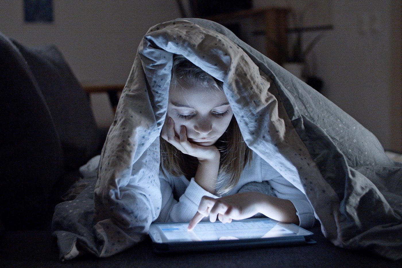 Niña bajo una manta con la luz de una tableta en la cara