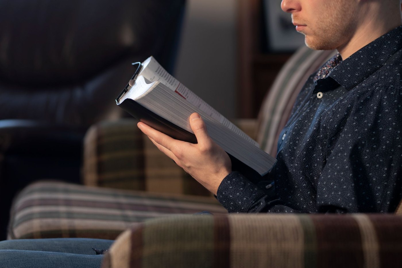 Un joven lee una Biblia gruesa sentado en un sillón, con luz cálida